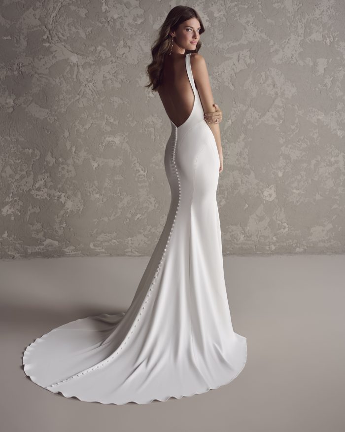 Bride wearing a sleek ivory crepe wedding dress with a dramatic low scoop back, covered button detailing trailing down the train, and a figure-hugging silhouette.