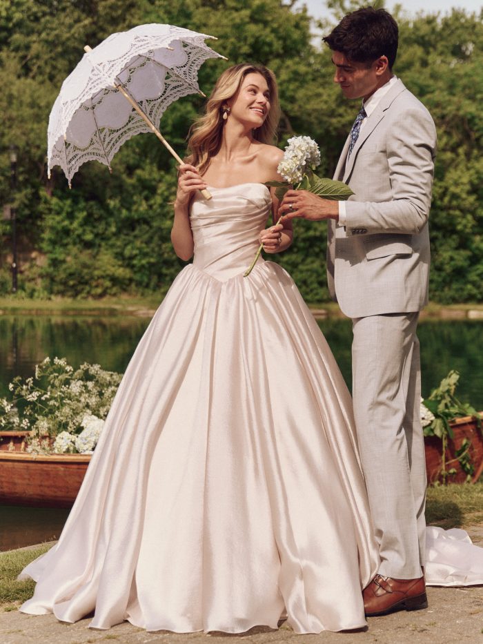 Bride wears Paris by Maggie Sottero—a strapless blush ballgown with a softly ruched bodice and voluminous skirt, holding a lace parasol while smiling at her partner in a garden setting.