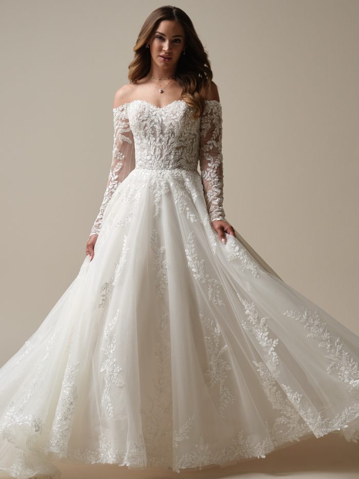 Bride wearing an off-the-shoulder A-line wedding dress with long illusion lace sleeves, a sheer embroidered bodice, and a full tulle skirt adorned with leafy lace appliqués.