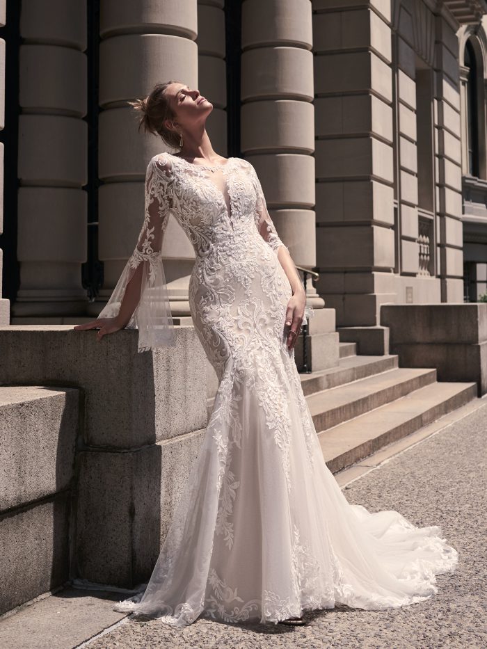 Front view of the Saige gown, showcasing the plunging illusion neckline, embroidered lace bodice, and dramatic bell sleeves, with the bride posing in sunlight against a historic building.