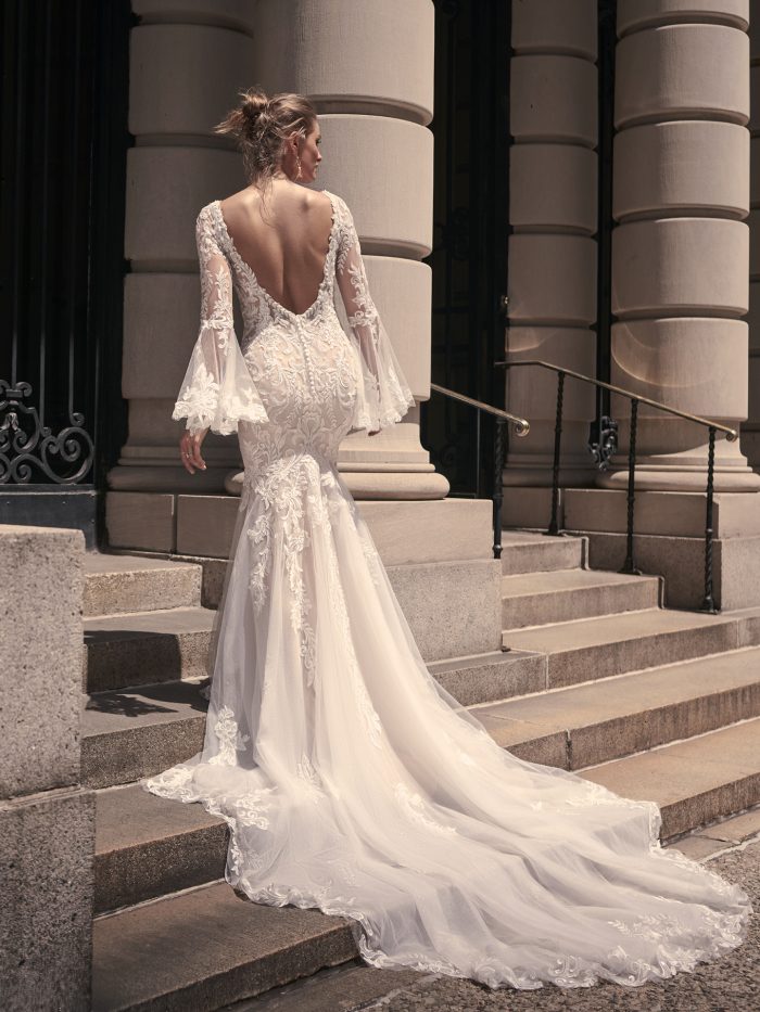 Back view of the Saige mermaid wedding dress, featuring a deep V-back, long illusion lace sleeves with flared cuffs, and a flowing embroidered tulle train as the bride ascends stone steps.