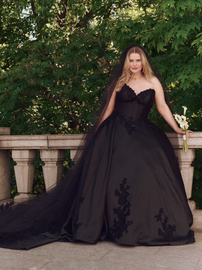 Front view of Maggie Sottero Serafina black ballgown wedding dress featuring illusion corset bodice, lace appliqués, and a long veil, styled with calla lilies.