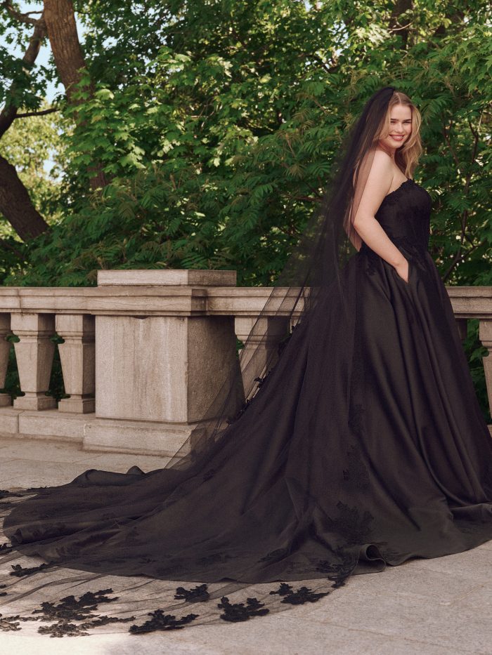 Model wearing Maggie Sottero Serafina black ballgown wedding dress with sweetheart neckline, full skirt, and dramatic lace-edged veil in an outdoor garden setting.