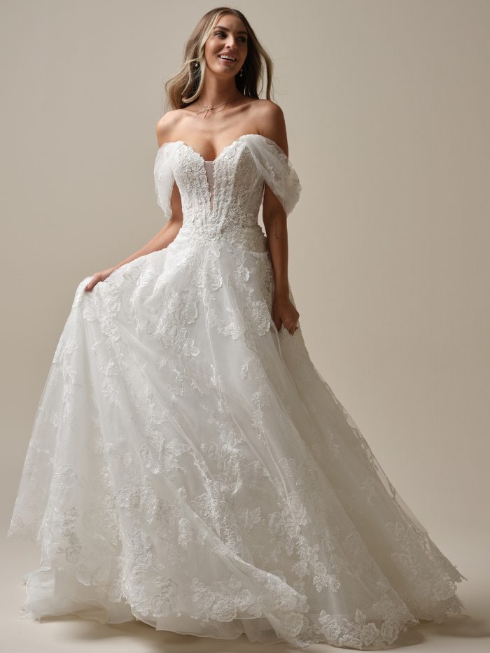 Bride wearing a romantic off-the-shoulder A-line wedding gown with floral lace embroidery, a sheer corset bodice, and flowing tulle skirt with a chapel-length train.