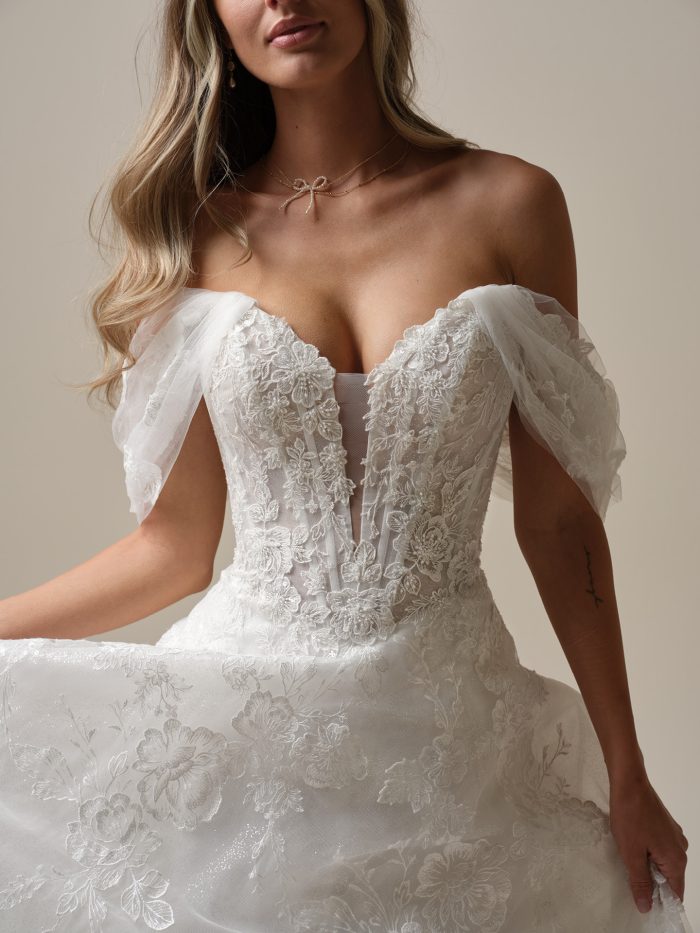 Close-up of an off-the-shoulder A-line wedding dress featuring a sheer lace bodice with exposed boning, floral embroidery, and soft tulle draped sleeves.