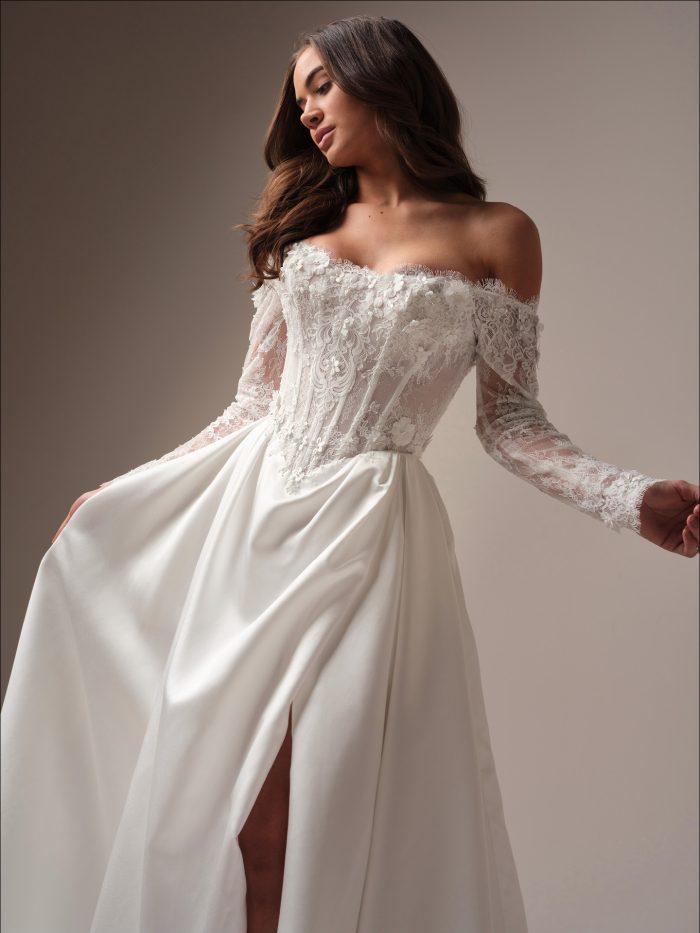 Bride wearing a regal off-the-shoulder A-line wedding dress with long illusion lace sleeves, a high slit, and a romantic corseted bodice adorned in 3-D floral lace.