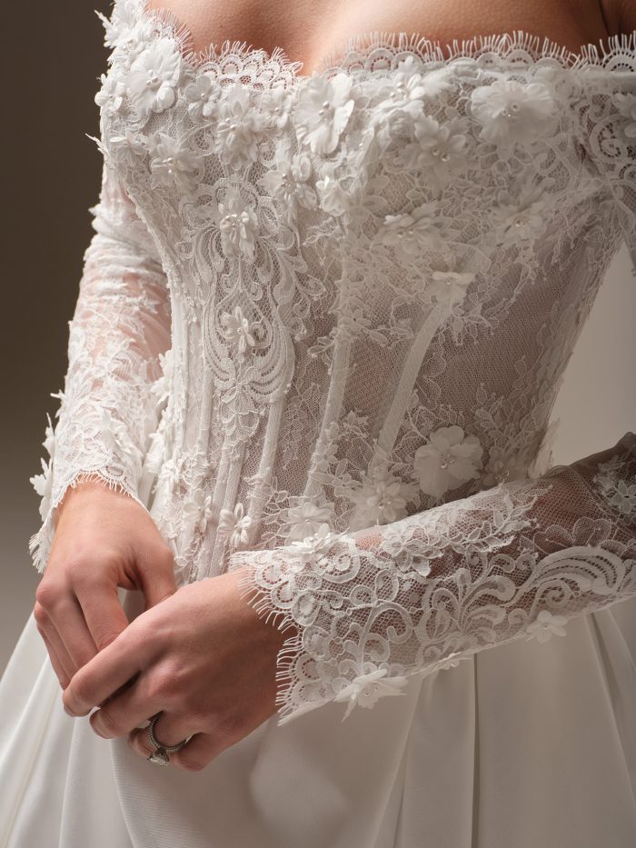 Close-up of a royal-inspired off-the-shoulder wedding dress featuring sheer lace sleeves, 3-D floral appliqués, and a structured corset bodice with pearl detailing.