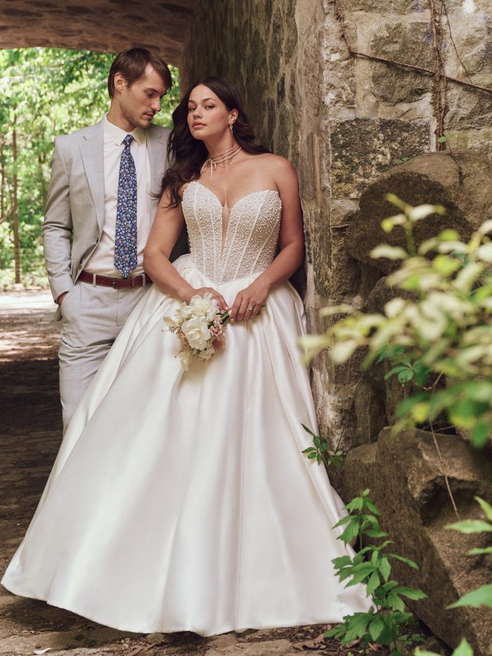 Bride in Maggie Sottero Tina ballgown wedding dress with pearl-embellished illusion bodice and full satin skirt, posing with groom in a romantic outdoor stone walkway.