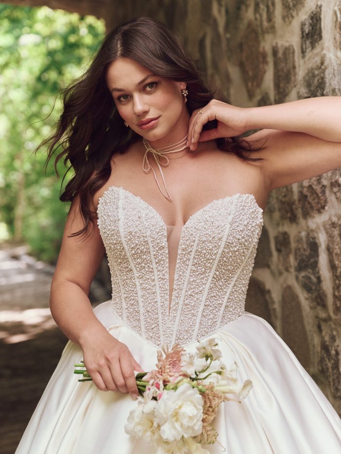 Close-up of bride wearing the Maggie Sottero Tina ballgown wedding dress featuring a pearl-embellished corset bodice with plunging neckline and satin skirt, holding a bouquet.