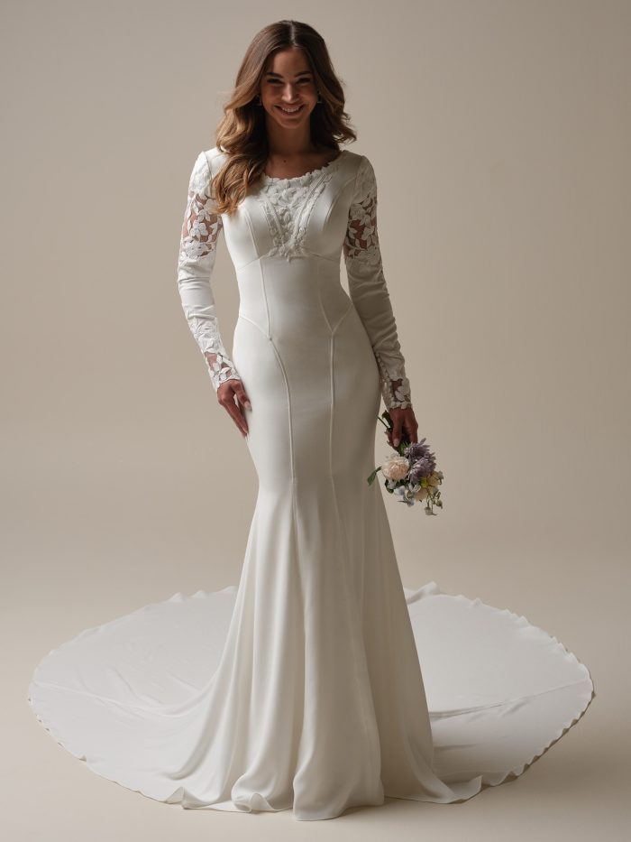 Bride wearing a crepe sheath wedding dress with a scalloped scoop neckline, lace-embellished illusion sleeves, and a dramatic chapel train, holding a soft pastel bouquet.