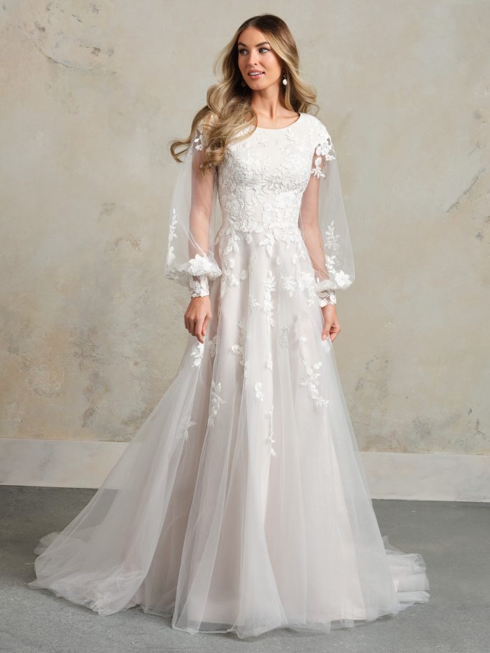 Bride wearing a modest A-line wedding dress with long sheer bishop sleeves, floral lace appliqués, a high illusion neckline, and soft tulle skirt for a romantic vintage look.