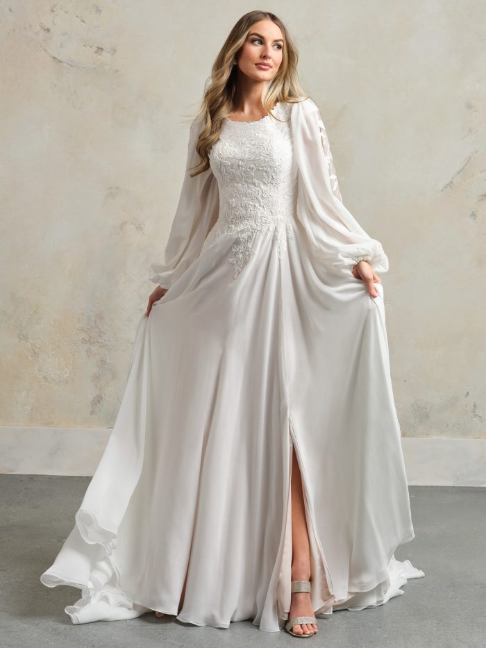 Bride wearing a romantic sheath wedding dress with long bishop sleeves, a lace bodice, soft chiffon skirt with front slit, and a high bateau neckline.