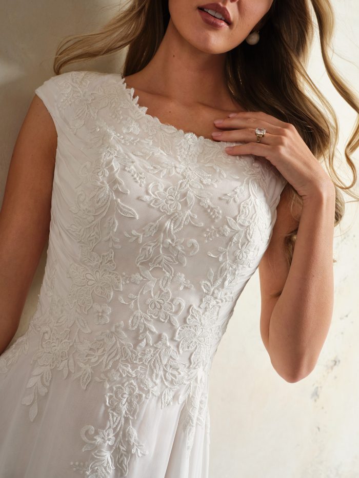 Close-up of a bridal gown bodice with cap sleeves, a bateau neckline, and delicate floral lace embroidery over sheer chiffon fabric.