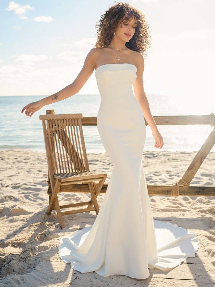 Front view of a strapless crepe wedding gown with a straight neckline and figure-hugging silhouette, styled with layered necklaces and loose curls on a beachside boardwalk.