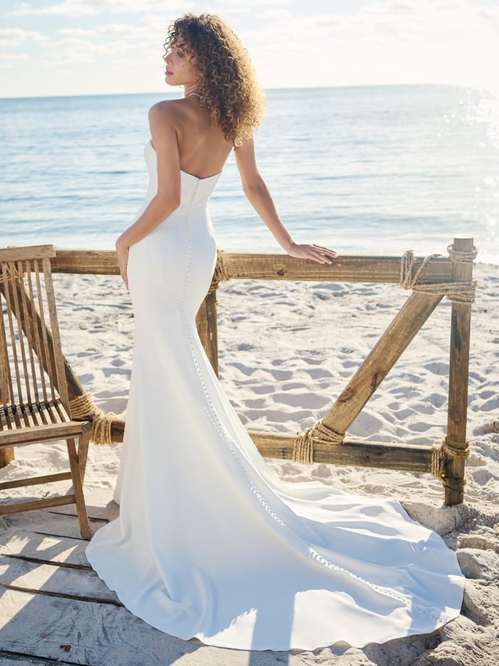 Bride wearing a strapless crepe sheath wedding dress with a soft scoop back, fabric-covered buttons trailing down the train, photographed on a sandy beach at sunset.