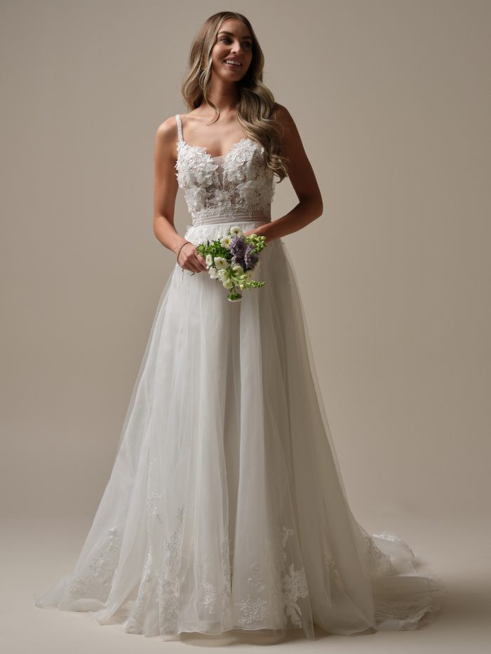 Bride wearing a romantic ballgown wedding dress with a sheer floral lace bodice, beaded spaghetti straps, and a soft tulle skirt adorned with scattered lace appliqués, holding a small pastel bouquet.