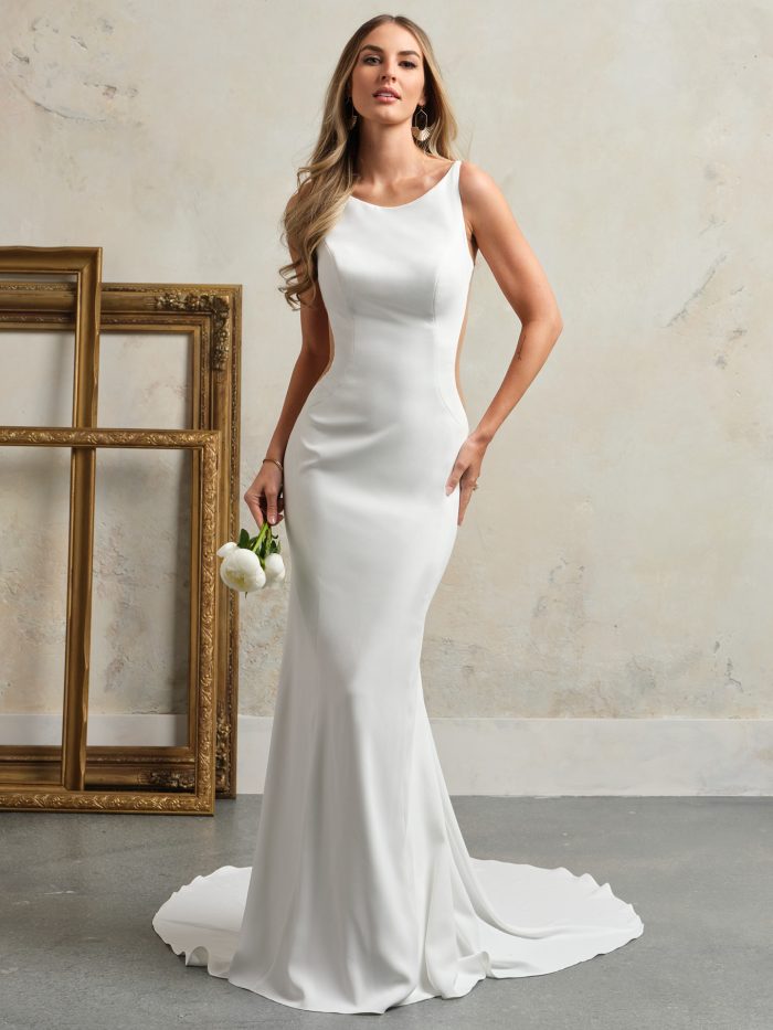 Bride wearing a minimalist sheath wedding dress with a high bateau neckline, side illusion cutouts, and soft crepe fabric, styled with loose curls and white roses.