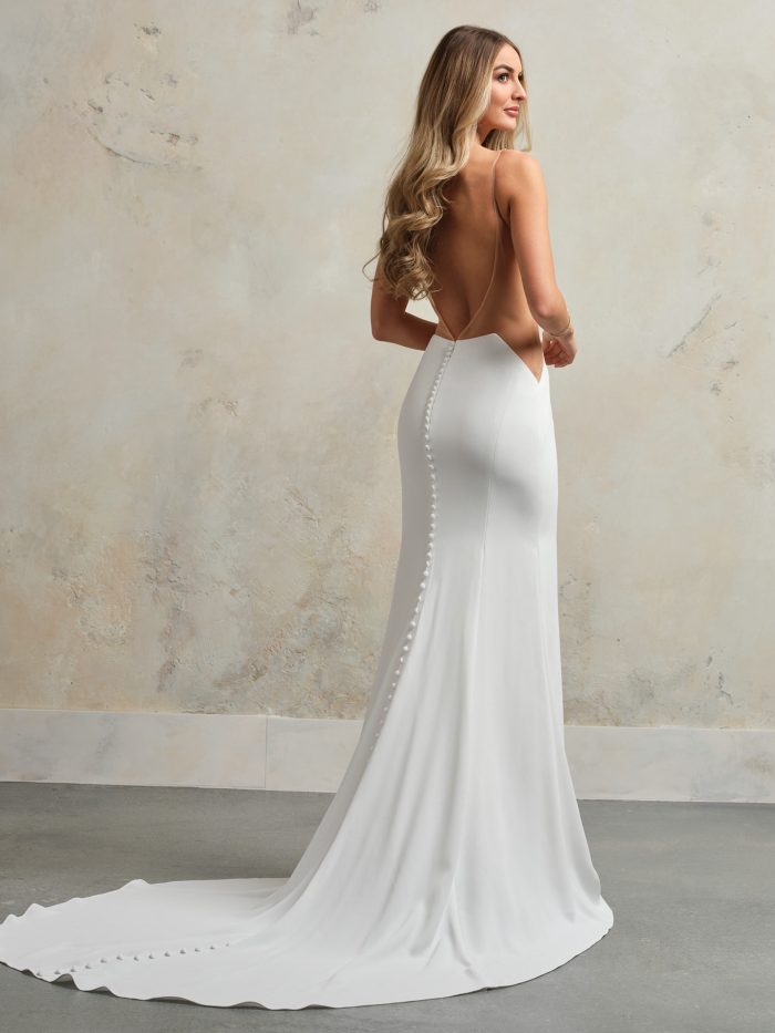 Back view of a modern crepe wedding gown with a dramatic low open back, thin spaghetti straps, and a row of buttons trailing down the train.