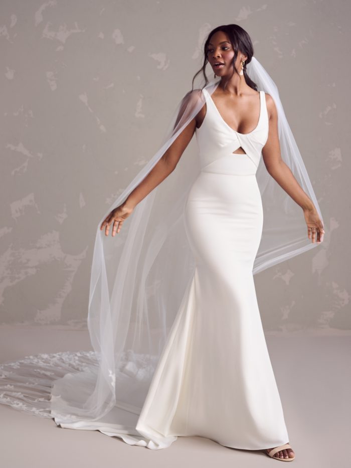 Front view of a sleek fit-and-flare wedding gown featuring a scoop neckline, subtle front cutout, and wide straps, styled with a long sheer veil and gold earrings.