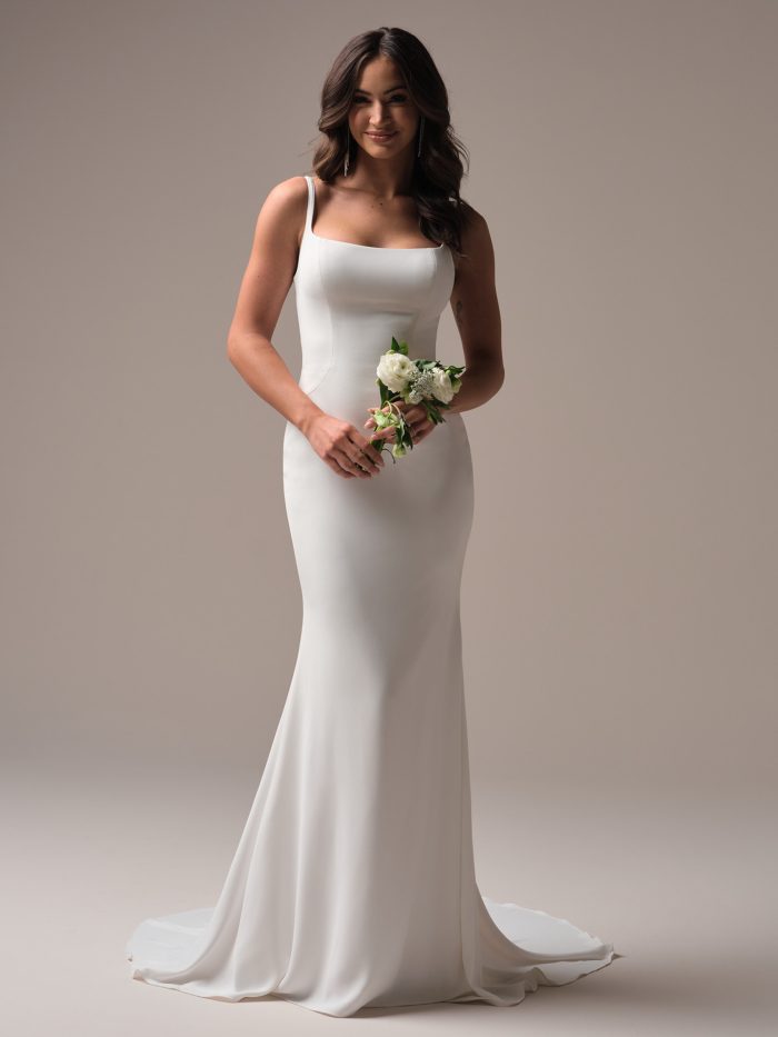 Front view of a minimalist sheath wedding gown with a square neckline and fitted bodice, styled with loose curls and a small white bouquet.