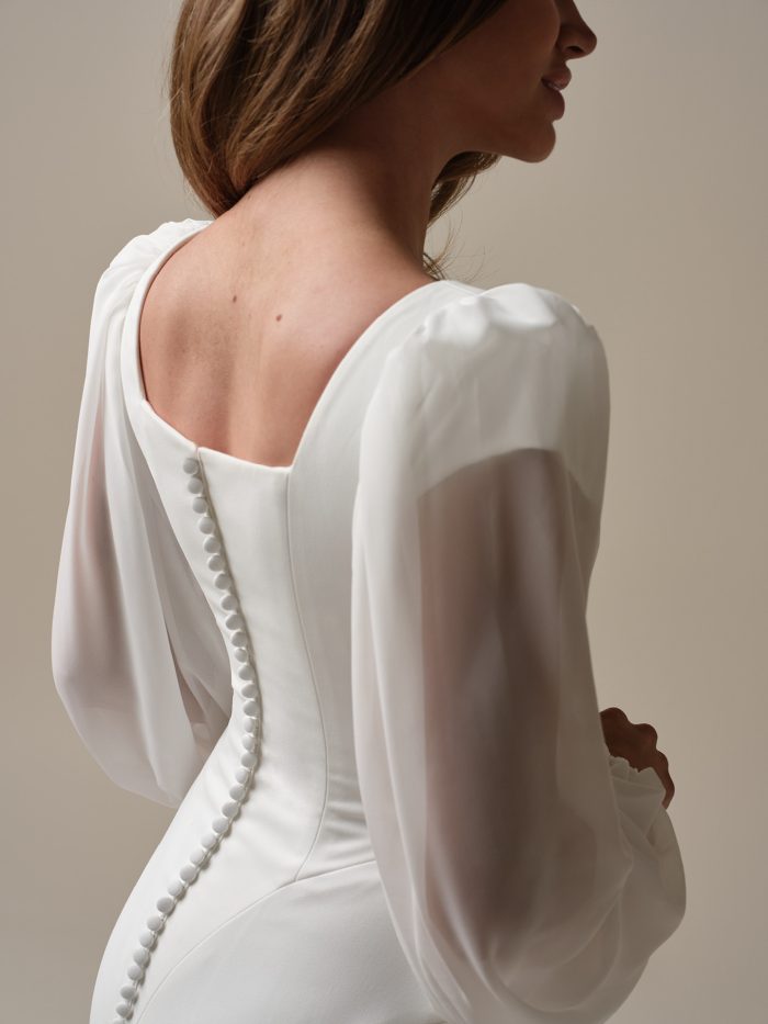 Back view of Jorie’s crepe sheath silhouette with fabric-covered buttons and puff sleeves—this modest wedding gown is minimalist perfection for the Gen Z bride who loves a soft statement.
