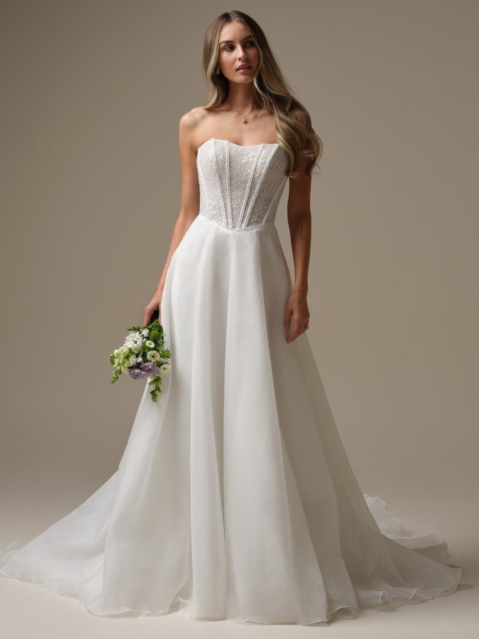 Bride wearing an A-line wedding dress with a sparkling, sequin-embellished strapless bodice and sheer boning detail, holding a bouquet with soft green and purple accents.