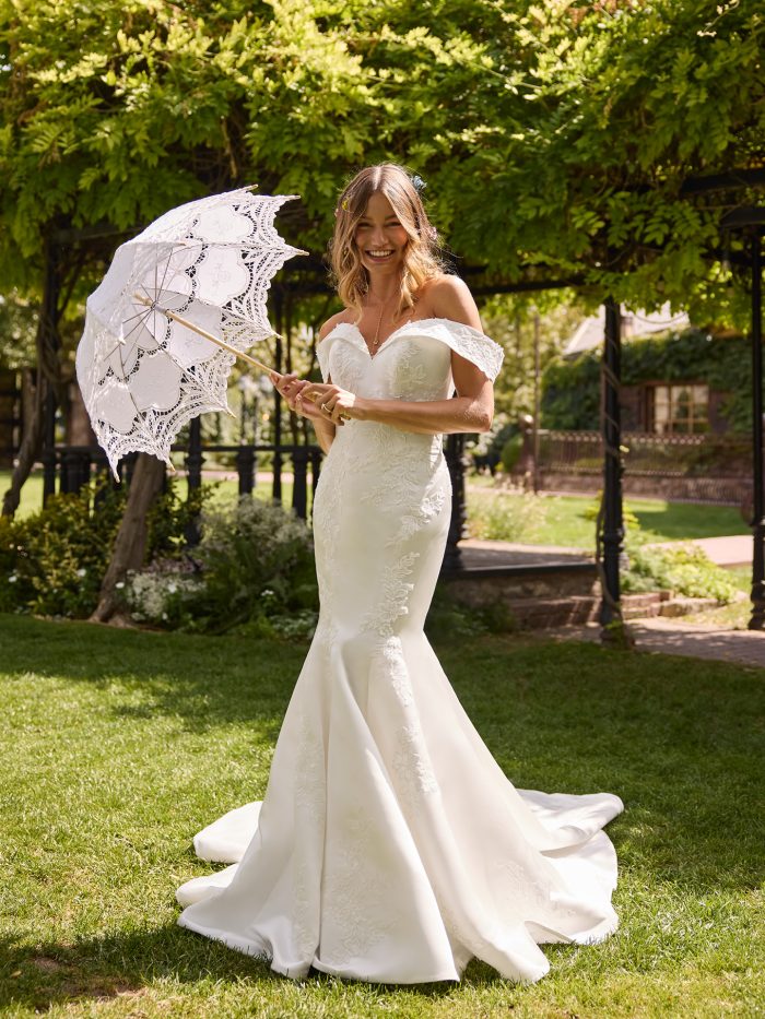 Bride wearing the Miley fit-and-flare wedding dress by Rebecca Ingram, featuring an off-the-shoulder neckline, floral lace detailing, and a softly flared skirt, holding a lace parasol in a garden setting.