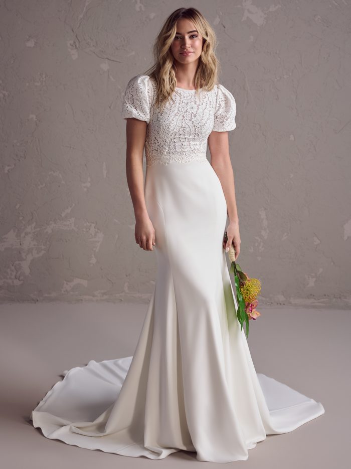 Bride wearing a fit-and-flare wedding dress with a puff-sleeve lace bodice, high scoop neckline, and sleek crepe skirt with a gentle train, holding a modern tropical bouquet.