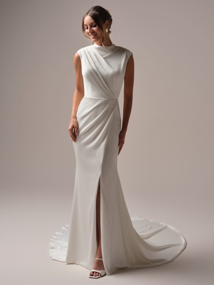 Front view of a modern satin wedding gown with an asymmetrical draped bodice, high neckline, and front skirt slit, styled with geometric earrings and a soft updo.