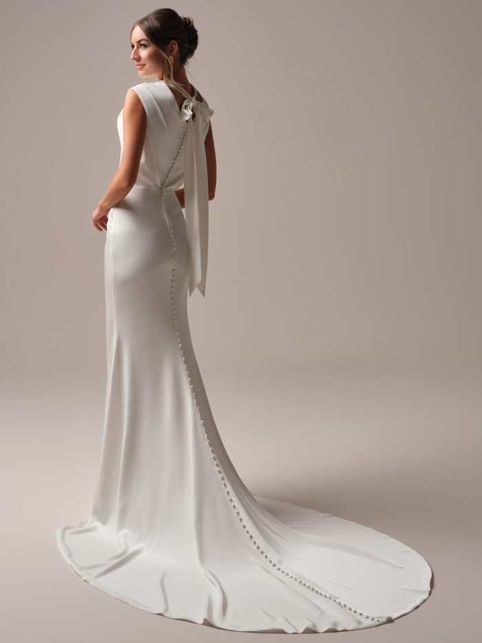 Bride wearing a satin sheath wedding dress with a high neckline, keyhole back accented by a dramatic bow, and fabric-covered buttons trailing down the train.