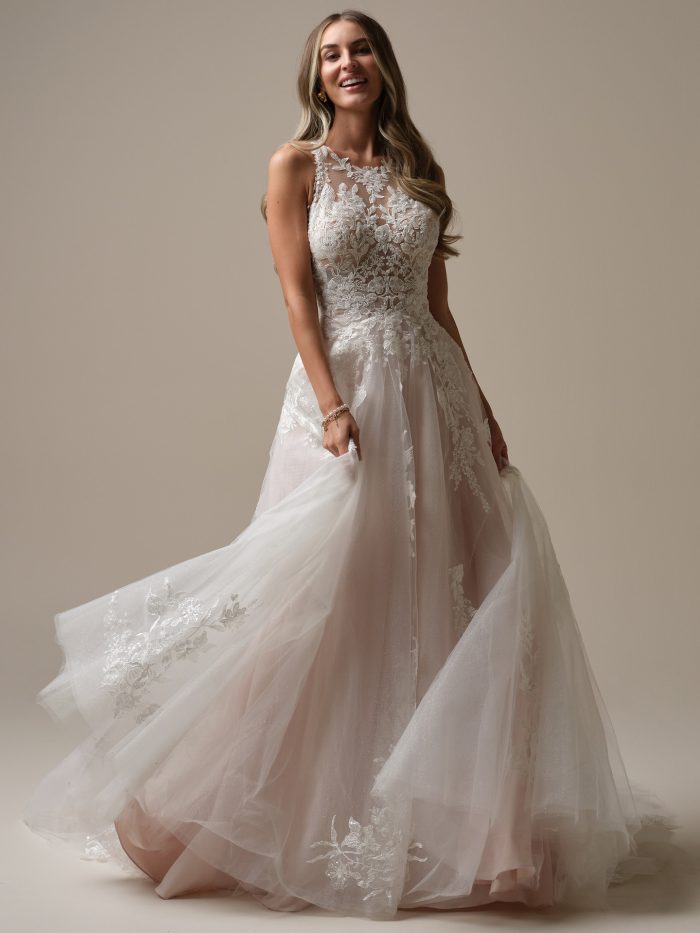 Full view of the Rickie gown in motion, featuring a floral illusion bodice and layered tulle A-line skirt with embroidered appliqués, as the bride twirls with a joyful expression in a studio setting.