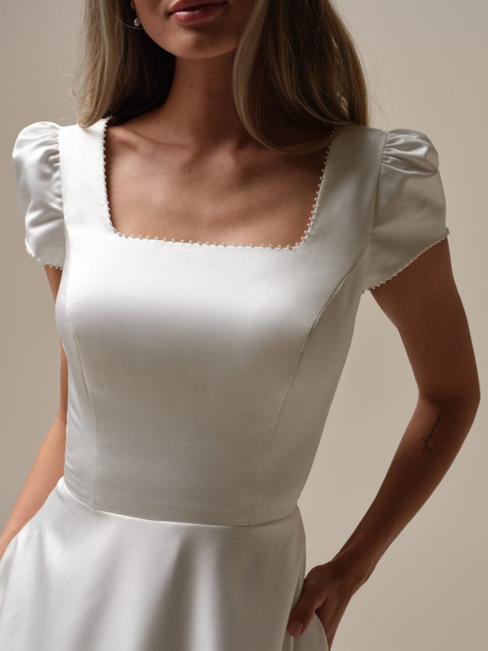 Close-up of a satin wedding dress bodice featuring a square neckline trimmed with pearl detailing and puff cap sleeves for a vintage-inspired touch.