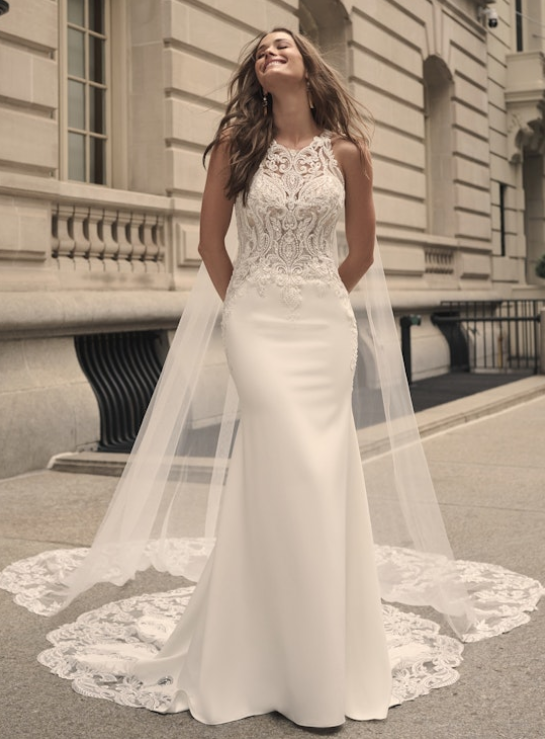 Front view of the Audrina sheath wedding gown, showcasing the illusion halter neckline, intricately beaded lace bodice, and lace-edged chapel-length train, worn by a smiling bride in an urban city setting.