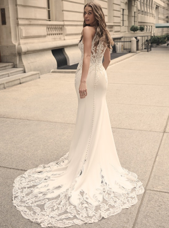 Back view of the Audrina wedding dress, highlighting the illusion lace back, fabric-covered buttons, and dramatic cutout lace train as the bride glances over her shoulder on a city sidewalk.