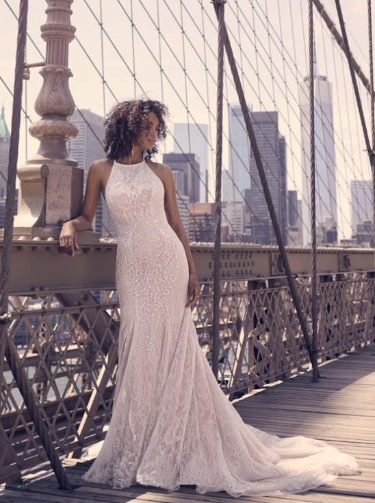 Front view of the Sloan gown, showcasing the high halter neckline, fitted lace silhouette, and subtle sparkle, as the bride poses against a bridge post overlooking the city skyline.