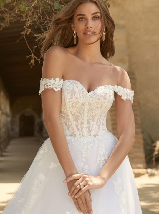 Bride wearing an off-the-shoulder ballgown wedding dress with a sheer corset bodice, sweetheart neckline, and lace appliqués over tulle for a romantic and feminine look.