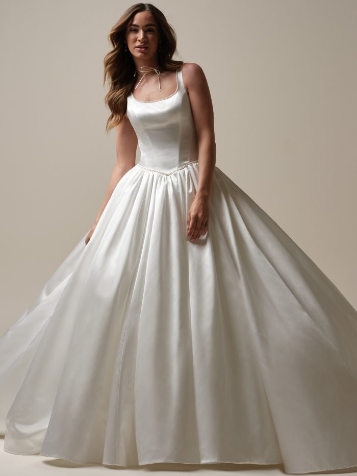 Bride wearing a classic satin ballgown wedding dress with a scoop neckline, pearl trim, and full pleated skirt, exuding timeless elegance.