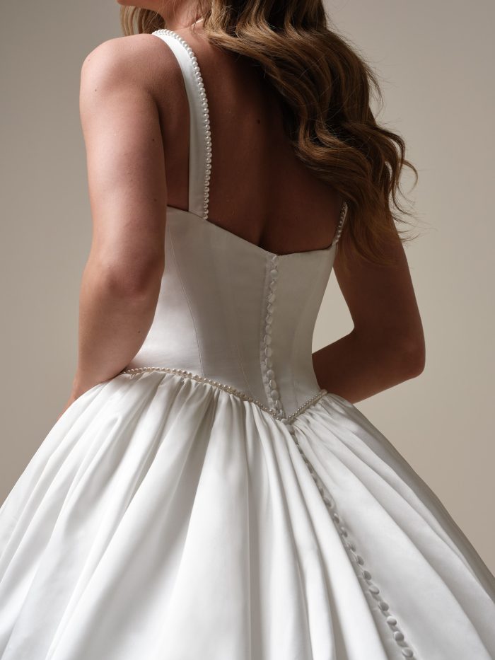 Back view of a bride wearing a satin ballgown wedding dress with a square back neckline, pearl-trimmed straps, and a row of satin buttons down the bodice and skirt.