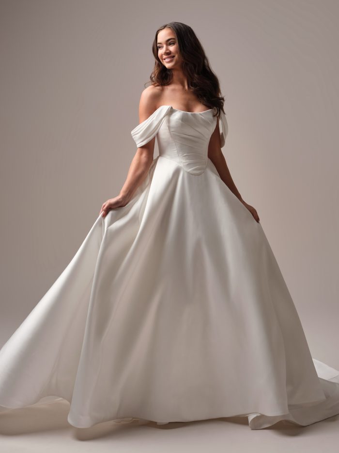 Full-length view of the Donovan wedding dress—a minimalist Mikado ballgown with off-the-shoulder sleeves, soft pleated bodice, and sweeping skirt; bride poses with a playful twirl and bright smile.