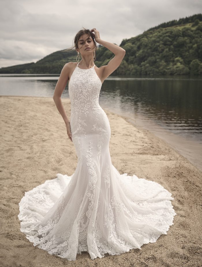 Front view of the Frost sheath wedding gown, featuring a high halter neckline, formfitting silhouette, and allover lace detailing, worn by a bride standing on a lakeside beach.