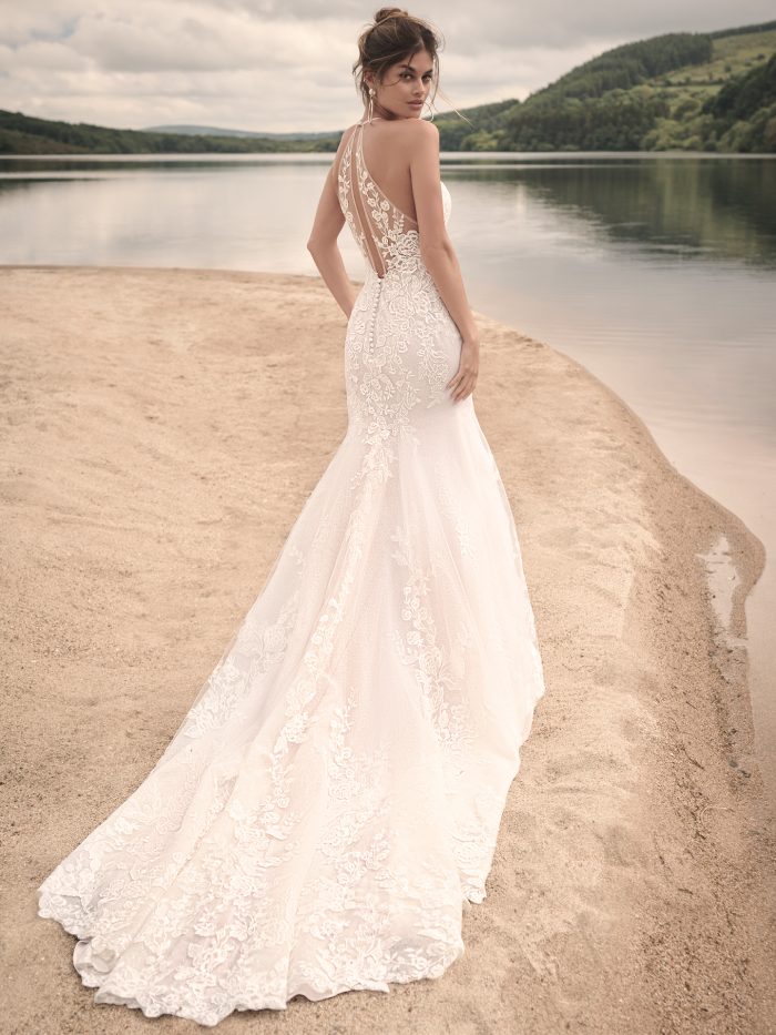 Back view of the Frost wedding dress, showcasing the illusion lace back with keyhole detail, fabric-covered buttons, and a lace train as the bride poses by the water’s edge.