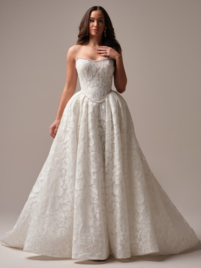 Model wearing Sottero and Midgley January ballgown wedding dress with strapless neckline, corset-style bodice, pearl-trimmed edges, and textured brocade-style floral fabric.