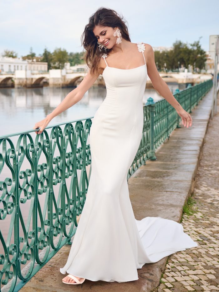 Bride wearing a minimalist sheath wedding gown with floral appliqué straps and a scoop neckline, smiling and posing playfully on a cobblestone walkway by the water.