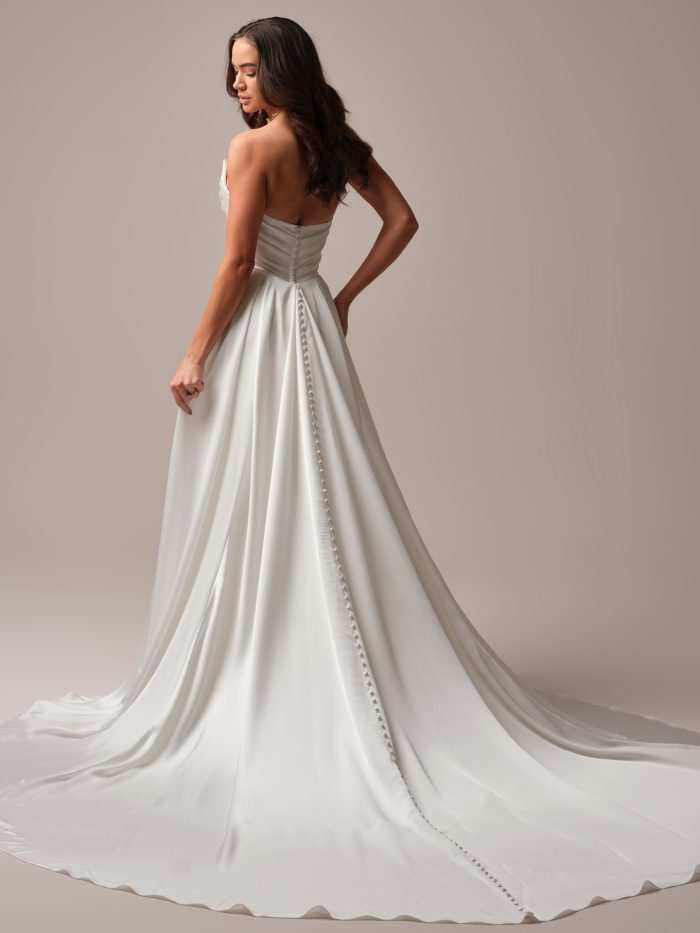Back view of a bride in a strapless satin sheath wedding dress with ruched bodice and a dramatic train accented by a line of satin-covered buttons.