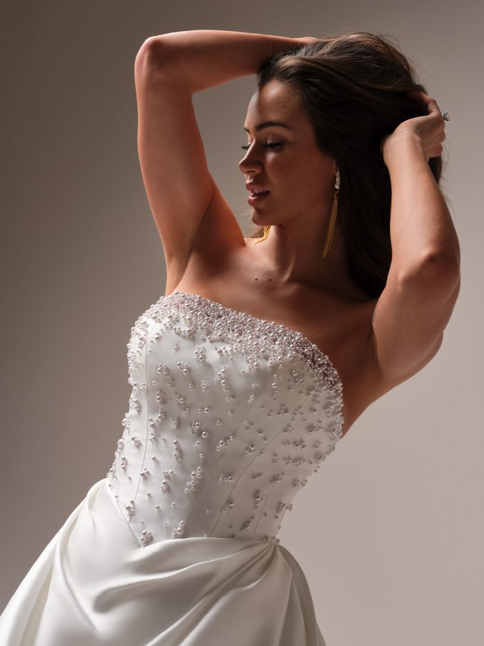 Bride wearing a strapless satin ballgown with a pearl-embellished bodice and soft ruching at the waist, striking a confident pose with hands in her hair.