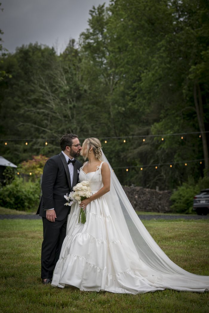 Bride and groom share a kiss outdoors beneath string lights, with the bride wearing a tiered satin ballgown and long veil while holding a white rose bouquet, and the groom dressed in a classic black tuxedo.
