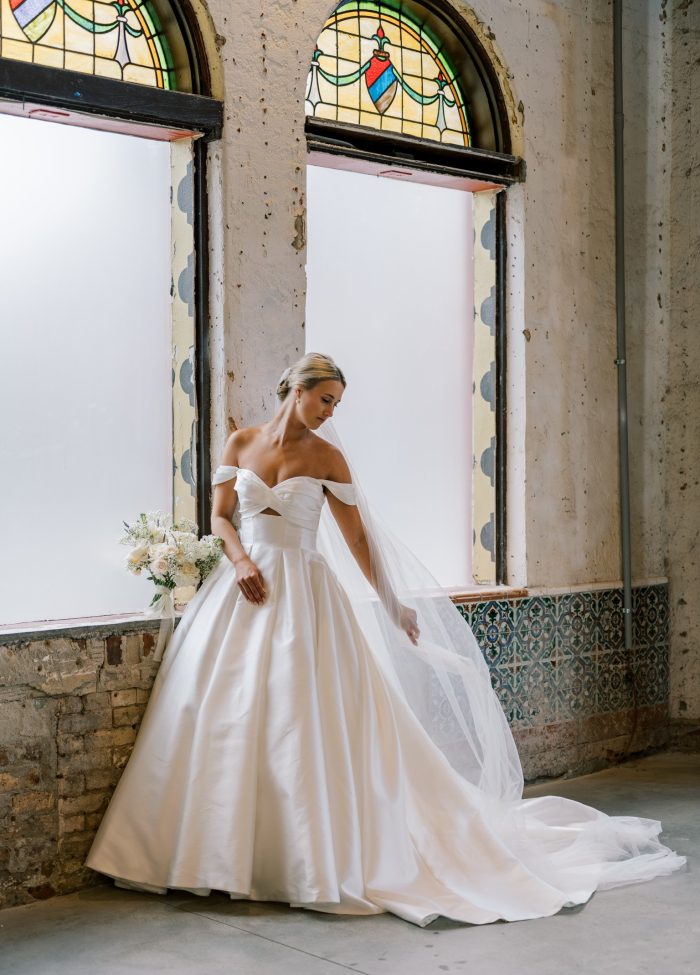 Bride wearing the Maggie Sottero Zinaida ballgown wedding dress with off-the-shoulder straps, bow detail bodice, and full satin skirt, posing by stained glass windows with a long veil and bouquet.