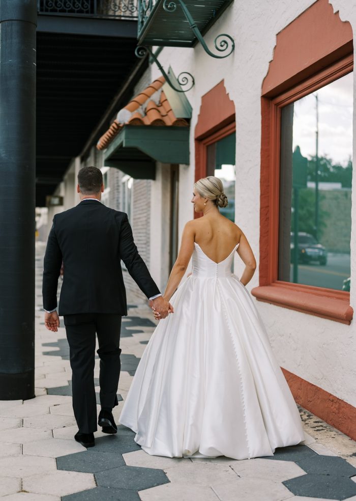 Bride wearing the Maggie Sottero Zinaida strapless satin ballgown with button-down back detail, walking hand-in-hand with groom in a classic outdoor city setting.