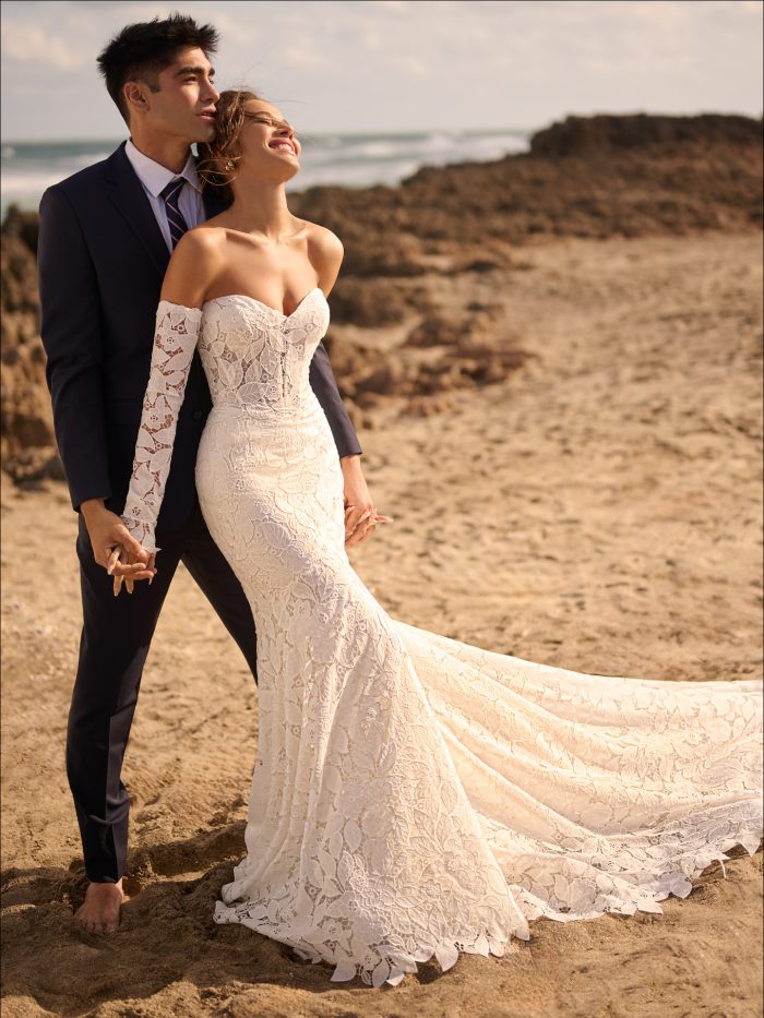 A bride wears the Maggie Sottero Dakota fit-and-flare wedding dress on the beach, featuring allover boho lace, a sweetheart neckline, and off-the-shoulder lace sleeves, with a flowing train trailing in the sand as she embraces her partner.