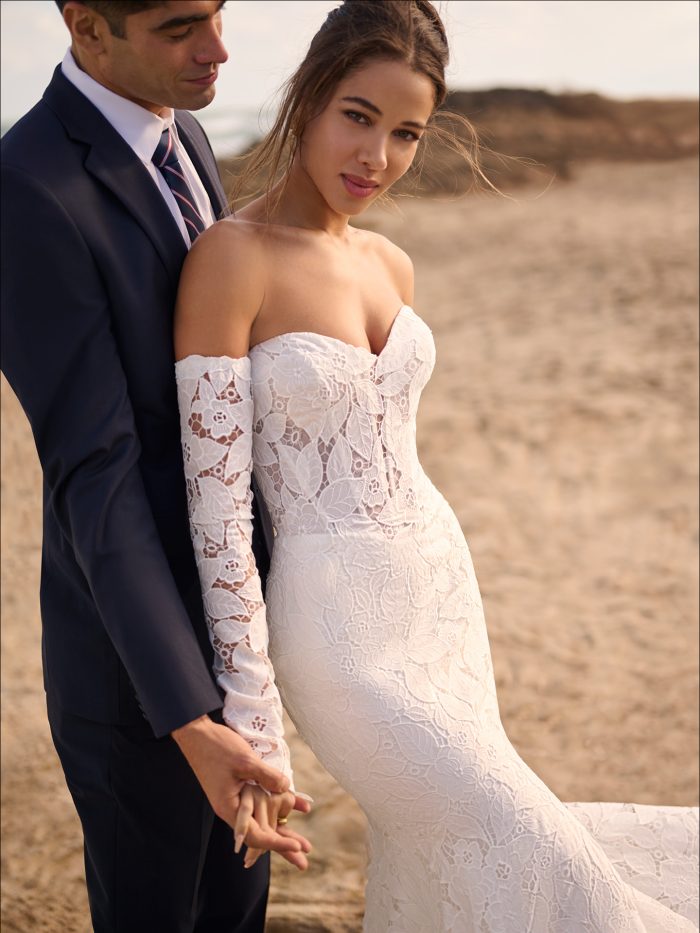 Close-up of the Maggie Sottero Dakota gown showing the intricate allover lace, off-the-shoulder sleeves, and fitted silhouette, as the bride and groom hold hands on the beach in a romantic sunset setting.